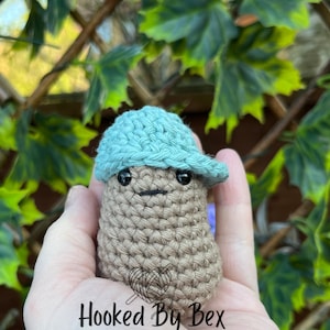 Op de afbeelding: Een gehaakte bruine aardappel met een lichtblauwe gehaakte hoed met rand. De aardappel heeft zwarte ogen en een kleine zwarte mond. De tekst "Hooked By Bex" is zichtbaar in het midden onderaan de afbeelding.
