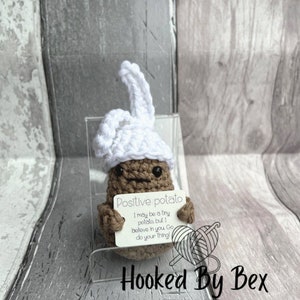 Może przedstawiać: Szydełkowa brązowa figurka ziemniaka w białej czapce kucharskiej, trzymająca znak z napisem "Positive potato". Znak zawiera tekst "I may be a tiny potato, but I believe in you. Go do your thing!" Figurka jest wystawiona na przezroczystym stojaku.