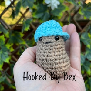 May include: A crocheted mushroom figure with a light blue cap. The mushroom is tan with black eyes and a small smile. The text "Hooked By Bex" is at the bottom of the image.