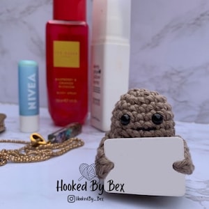 Op de afbeelding: Een gehaakt bruin personage dat een leeg wit bord vasthoudt. Het personage heeft een glimlachend gezicht met zwarte ogen. Het bord is leeg en klaar voor een bericht.