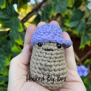 May include: A crocheted brown potato wearing a purple crocheted hat with a black face and a mustache. The text "Hooked By Bex" is visible on the potato.