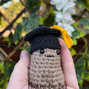 May include: A crocheted peanut wearing a black graduation cap with a yellow tassel. The peanut has a brown body and black eyes. The text "Hooked By Bex" is visible on the peanut's body.