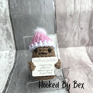 Può includere: Una patata all'uncinetto con un cappello rosa e bianco tiene un cartello che recita "Positive potato I may be a tiny potato, but I believe in you Go do your thing!" Il cartello è su uno sfondo bianco con le parole "Hooked By Bex" in basso.