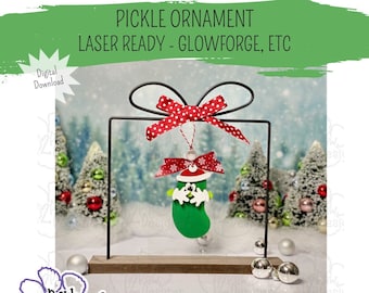 May include: A green pickle ornament with a red Santa hat and white trim. The ornament is hanging from a black metal stand with a red and white polka dot bow. The stand is on a wooden base with two silver balls.