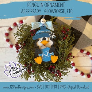 Pode incluir: Um enfeite de pinguim azul com um chap&eacute;u azul e um floco de neve no topo. O pinguim est&aacute; segurando um enfeite azul e branco listrado. O enfeite &eacute; cercado por uma grinalda verde com bagas vermelhas. O enfeite &eacute; etiquetado com "2022".