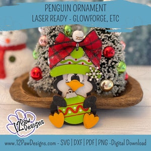 Pode incluir: Um ornamento de pinguim de madeira com um la&ccedil;o xadrez vermelho e preto, um chap&eacute;u verde com bolinhas brancas e uma bola de Natal verde e branca. O ornamento est&aacute; pronto para ser cortado com um cortador a laser.