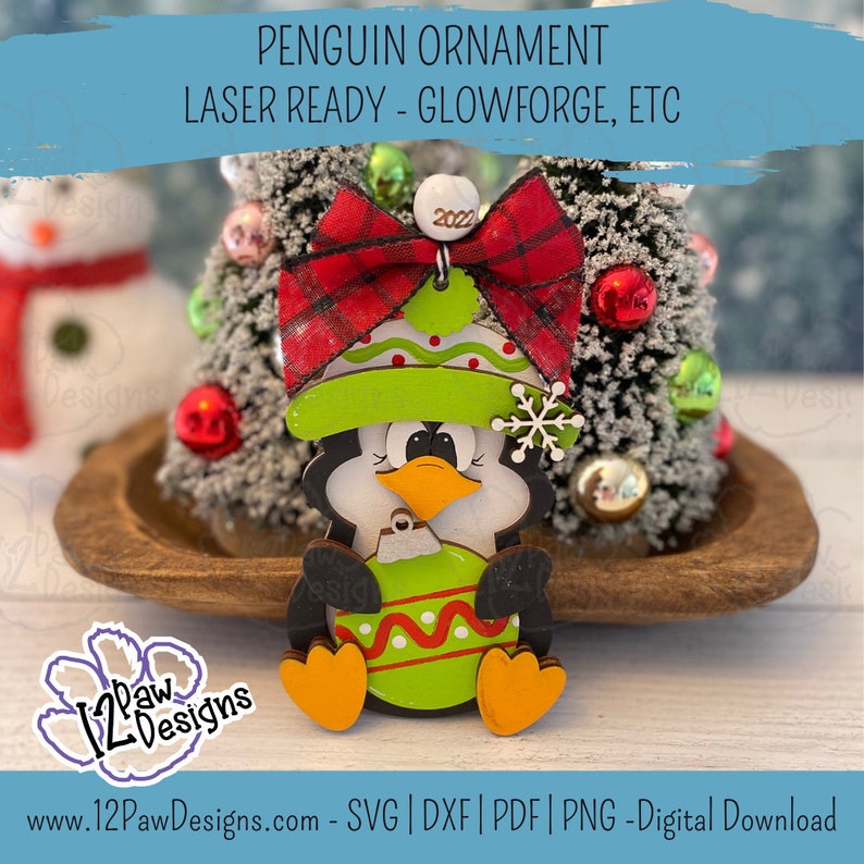Pode incluir: Um ornamento de pinguim de madeira com um la&ccedil;o xadrez vermelho e preto, um chap&eacute;u verde com bolinhas brancas e uma bola de Natal verde e branca. O ornamento est&aacute; pronto para ser cortado com um cortador a laser.