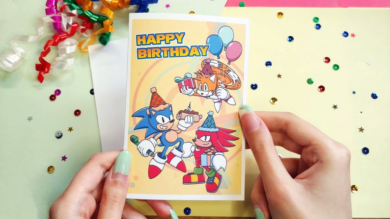 K&ouml;nnte beinhalten: Eine gelbe Geburtstagskarte mit den Worten "Happy Birthday" in Regenbogen-Schrift. Die Karte zeigt eine Comic-Illustration von Sonic the Hedgehog, Tails und Knuckles, die einen Geburtstag mit Kuchen, Ballons und Geschenken feiern.