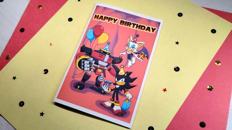 Sonic birthday card laying on table that says Happy Birthday at the top in a colorful Sonic themed font. Image has Team Dark (Shadow, Rouge, and Omega) holding gifts.