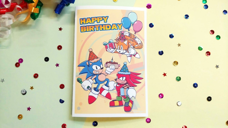 K&ouml;nnte beinhalten: Eine Geburtstagskarte mit Sonic the Hedgehog, Tails und Knuckles. Die Karte hat einen gelben Hintergrund mit einem Regenbogen-Wirbelmuster. Die Charaktere tragen Partyh&uuml;te und halten Geschenke. Der Text "HAPPY BIRTHDAY" ist oben auf der Karte in Schwarz gedruckt.