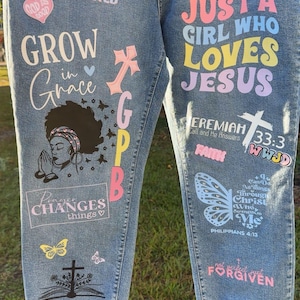 May include: Light blue denim jeans with various colorful graphics and text. The text includes phrases like "Bloom Where You Are Planted," "Just a Girl Who Loves Jesus," and Bible verses. The jeans feature a distressed hem and a variety of religious symbols.