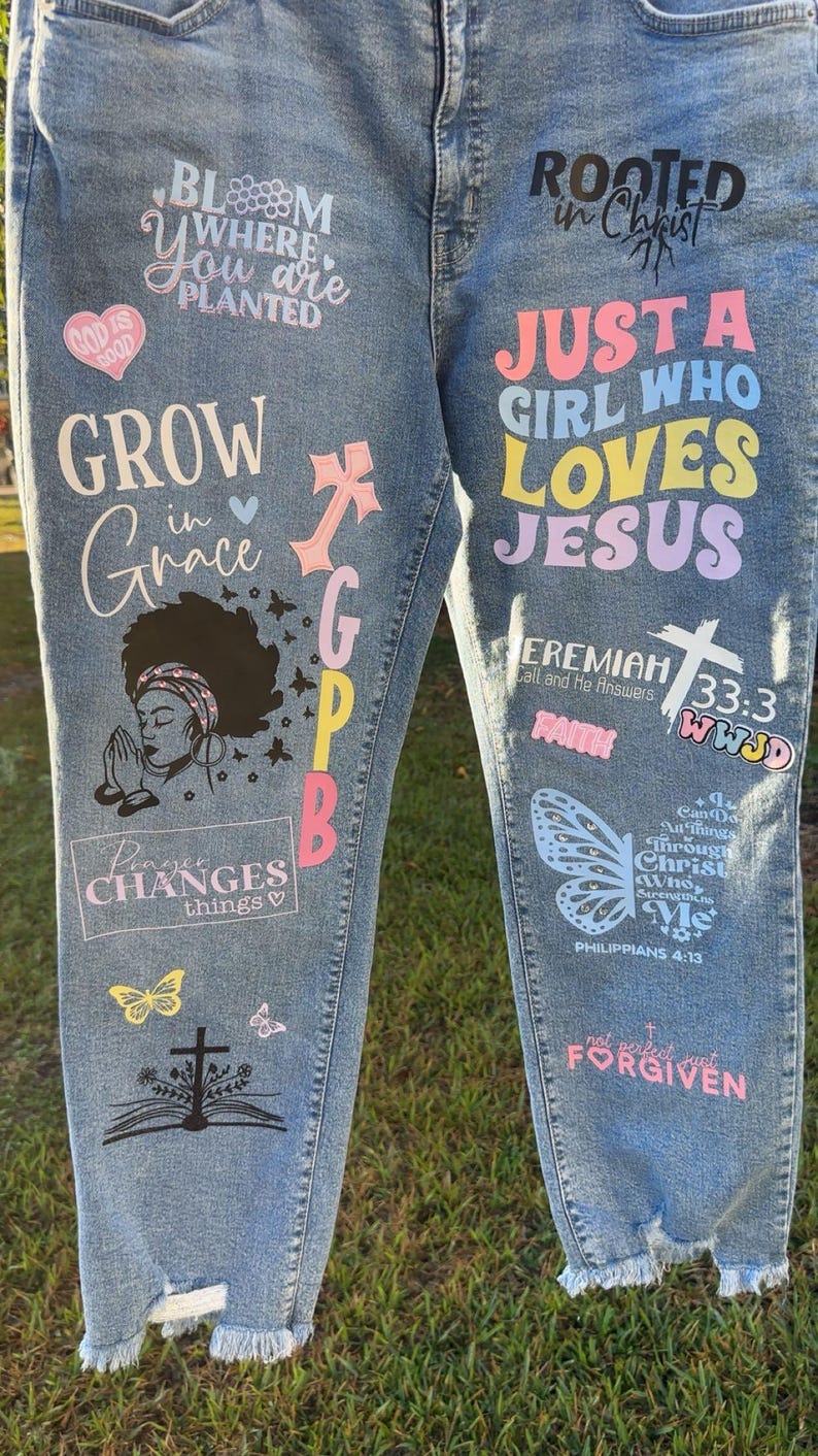 May include: Light blue denim jeans with various colorful graphics and text. The text includes phrases like "Bloom Where You Are Planted," "Just a Girl Who Loves Jesus," and Bible verses. The jeans feature a distressed hem and a variety of religious symbols.