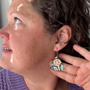 Puede incluir: Pendientes triangulares con un diseño floral negro, dorado, rojo y verde. Los pendientes cuelgan de ganchos dorados. Un accesorio único.