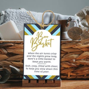 May include: A woven basket filled with a gray towel, white soap, and a small metal container. A decorative tag with the words "B+++ Basket" and a poem is displayed in front of the basket. The basket is on a wooden surface.