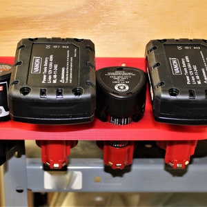 May include: A red plastic wall-mounted organizer with four slots for holding power tool batteries. The organizer holds two black batteries labeled "VANON" and two smaller batteries, one labeled "BOSCH".