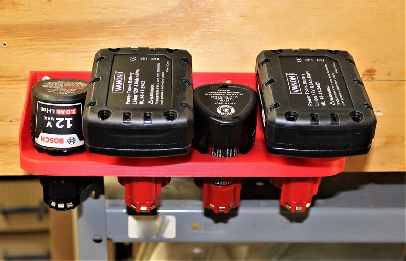 May include: A red plastic wall-mounted organizer with four slots for holding power tool batteries. The organizer holds two black batteries labeled "VANON" and two smaller batteries, one labeled "BOSCH".