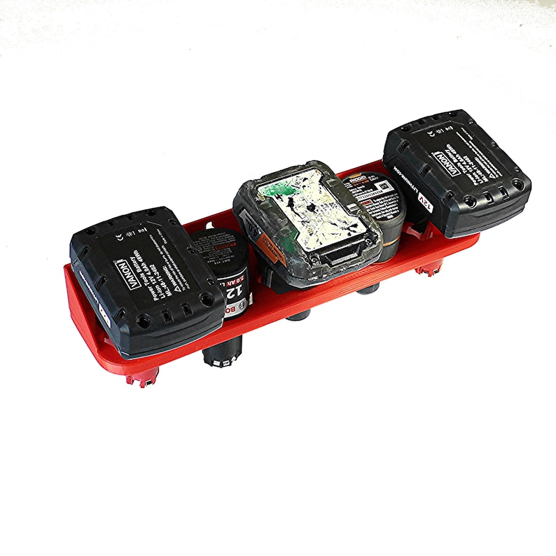 Puede incluir: Un estante de almacenamiento de bater&iacute;as de pl&aacute;stico rojo con cuatro ranuras. Tres ranuras est&aacute;n llenas de bater&iacute;as de herramientas el&eacute;ctricas negras y verdes. Las bater&iacute;as tienen el texto "VANON" y "12V" impreso en ellas.