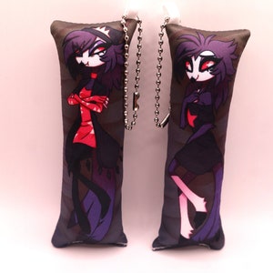 May include: Two plush keychain pillows featuring a cartoon character with black hair, red eyes, and a black and red outfit. The character is depicted in two different poses, one with arms crossed and the other with one hand on the hip. The pillows are attached to silver chains.