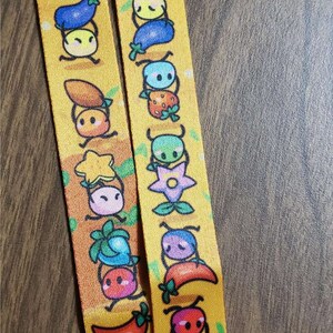May include: Two yellow lanyards with a repeating pattern of colorful cartoon characters holding various fruits and vegetables. The characters are all smiling and have large eyes. The lanyards are made of a soft, woven fabric.