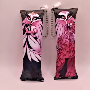 May include: Two rectangular plush keychains featuring a character with a pink and white color scheme, wearing a dress. The keychains have a dark brown top and a black bottom. Each keychain has a silver chain attached.