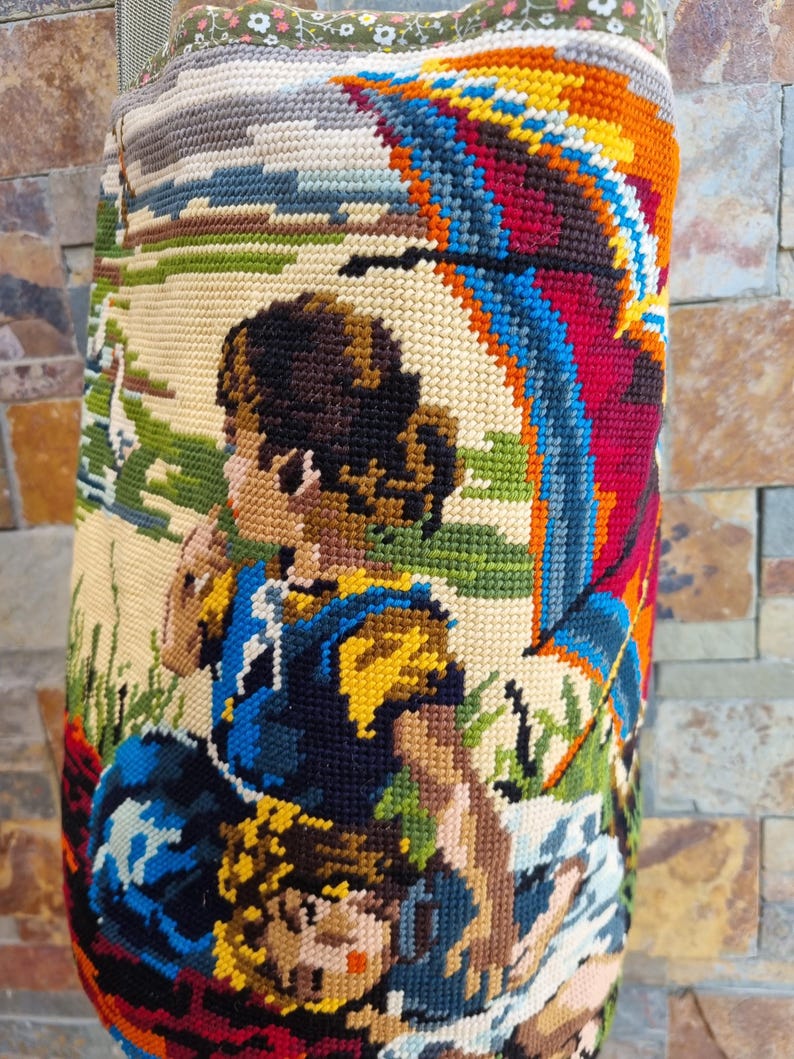 Op de afbeelding: Een tapijtzak met een kleurrijke sc&egrave;ne van figuren onder een regenboogkleurige paraplu. De tas heeft een gedetailleerd kruissteekontwerp met verschillende kleuren, waaronder blauw, geel, rood en groen. De bovenkant van de tas heeft een bloemenpatroon.