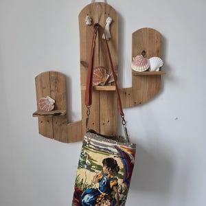 Op de afbeelding: Een rustieke, houten cactusvormige plank met bevestigde schelpen. Een tapijttas met een kleurrijke sc&egrave;ne van figuren en een landschap hangt aan de plank. De tas heeft een bruine leren riem.