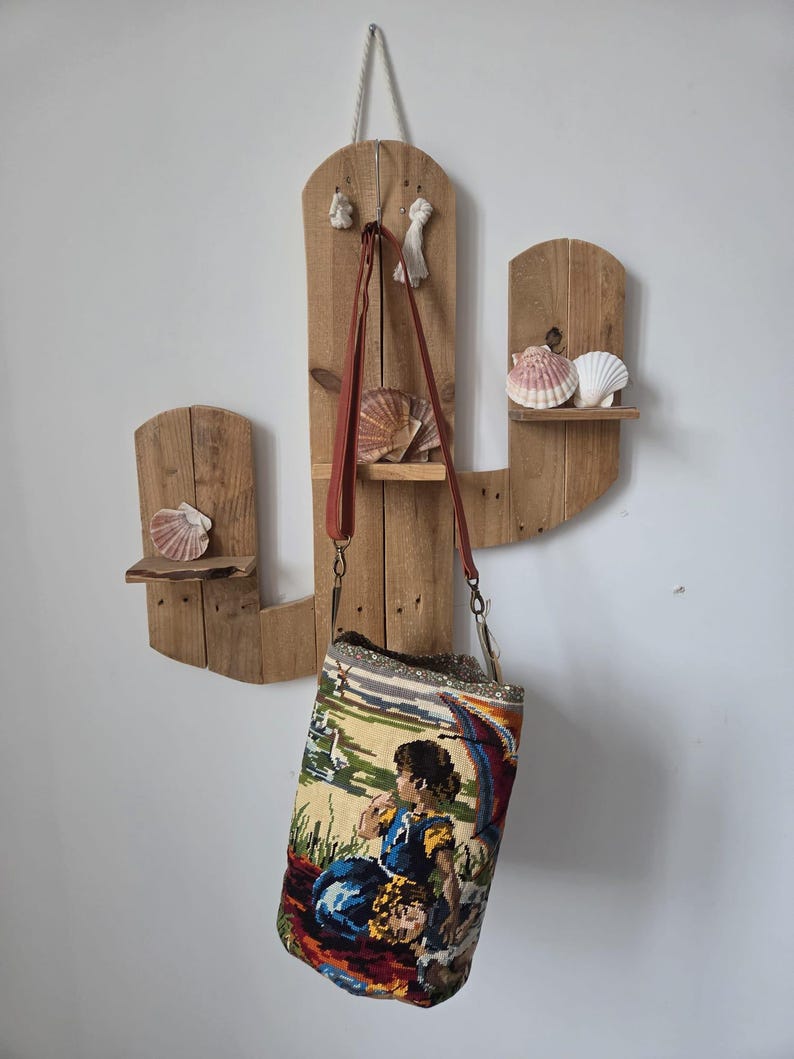 Op de afbeelding: Een rustieke, houten cactusvormige plank met bevestigde schelpen. Een tapijttas met een kleurrijke sc&egrave;ne van figuren en een landschap hangt aan de plank. De tas heeft een bruine leren riem.