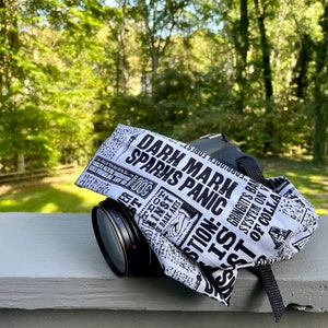 May include: A black camera with a lens cover featuring a black and white newspaper print design. The text on the cover reads "DARK MARK SPARKS PANIC" and other phrases. The camera is on a gray surface with a green background.