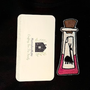 May include: A pink and white embroidered patch of a potion bottle with a cork stopper and a silhouette of a llama inside. The patch has the text "MamaAndKate Crafting For The Family" on a white card behind it.