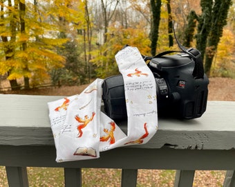 May include: A black Canon EOS Rebel T5i DSLR camera with a white camera strap. The strap features a repeating pattern of orange phoenixes, open books, stars, and script on a white background. The camera is resting on a gray surface.