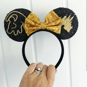 May include: Black mouse ears with a gold glitter bow. One ear features a gold silhouette of Simba from The Lion King, the other ear features a gold silhouette of Cinderella's castle.