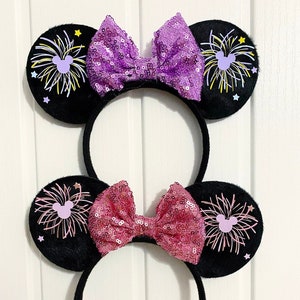 May include: Two pairs of black mouse ears with a purple and pink bow. The ears have a firework design with a pink Mickey Mouse silhouette.