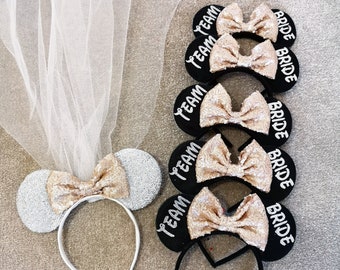 Puede incluir: Conjunto de seis orejas de ratón negras con diadema y lazos de purpurina dorada. Las diademas tienen las palabras "Team" y "Bride" escritas en ellas.