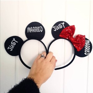 Puede incluir: Un par de orejas de Mickey Mouse negras con un lazo rojo de lentejuelas. Las orejas est&aacute;n decoradas con las palabras "Just Married" en purpurina plateada.