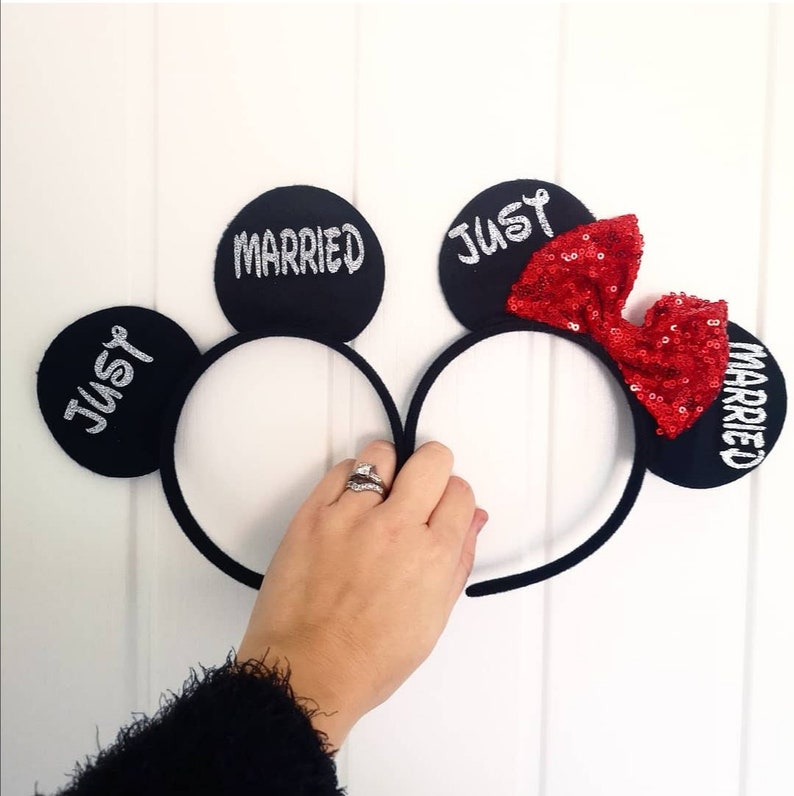 Puede incluir: Un par de orejas de Mickey Mouse negras con un lazo rojo de lentejuelas. Las orejas est&aacute;n decoradas con las palabras "Just Married" en purpurina plateada.