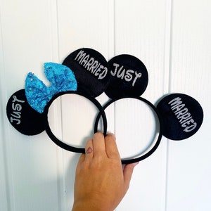 Puede incluir: Un par de orejas de rat&oacute;n negras con un lazo de lentejuelas azules. Las orejas est&aacute;n decoradas con letras plateadas brillantes que dicen "Just Married" en una oreja y "Married Just" en la otra.
