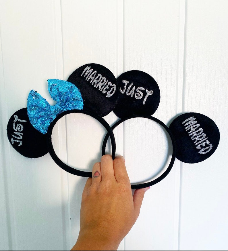 Puede incluir: Un par de orejas de rat&oacute;n negras con un lazo de lentejuelas azules. Las orejas est&aacute;n decoradas con letras plateadas brillantes que dicen "Just Married" en una oreja y "Married Just" en la otra.