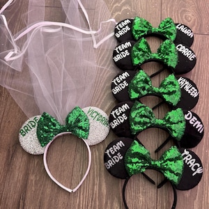 May include: A collection of bridal party headbands and a veil. The headbands feature black or silver ears with green sequin bows. Names like "Laura", "Corrie", and "Victoria" are printed on the ears. The veil is white.