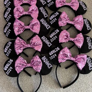 May include: A set of black mouse ears with pink sequined bows. The ears are decorated with the text "RADIANCE" and "WORLDS 2024".