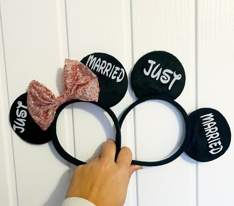 Puede incluir: Orejas de Mickey Mouse negras con un lazo rosa y las palabras "Just Married" en cada oreja.