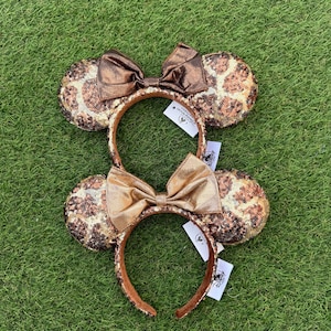 Puede incluir: Dos diademas con orejas redondas cubiertas de lentejuelas marrones y doradas con estampado de leopardo. Cada diadema tiene un lazo metálico en el centro, uno en tono bronce oscuro y otro en tono dorado claro, con etiquetas de Disney Resort.