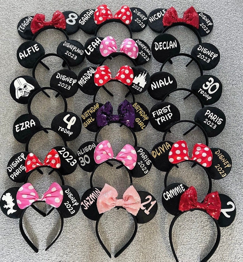 May include: A collection of 15 black mouse ear headbands with various personalized designs. The headbands feature different colored bows, including red, pink, purple, and sequins. The designs include names, numbers, and phrases like "Disney 2023", "Birthday Girl", and "First Trip".