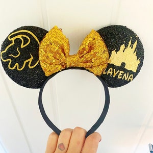 May include: Black mouse ears with a gold bow, a gold lion paw print on one ear, and a gold castle silhouette on the other ear. The text "LAVENA" is written on the castle ear.