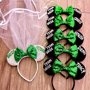 Peut inclure: Une collection d'accessoires de mariage, comprenant un voile blanc et plusieurs bandeaux à oreilles de souris à paillettes. Les bandeaux présentent des nœuds verts et le texte "Bride" et "Team Bride", ainsi que des noms comme "Laura" et "Tracy".