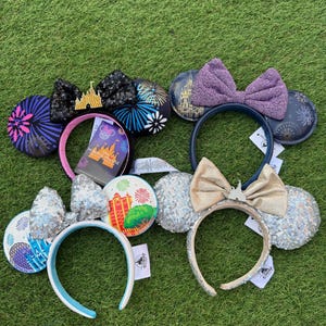 May include: A collection of Minnie Mouse ear headbands with various designs. Some feature sequined ears and bows, while others showcase fireworks, castles, and park icons. The headbands have different colored bands, including pink, blue, and gold, and are displayed on a green surface.