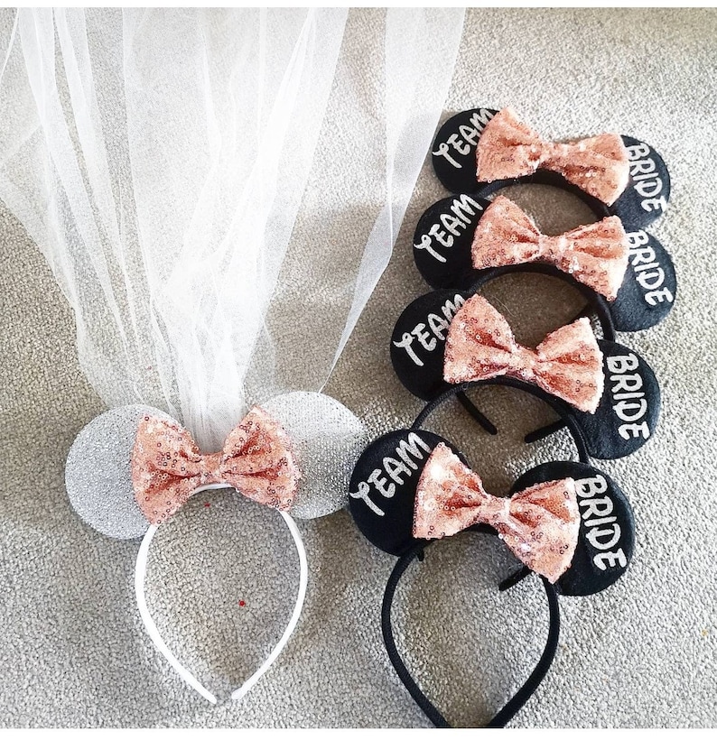 Op de afbeelding: Vier zwarte haarbanden met roze pailletten strikken en witte tekst. Een haarband heeft zilveren pailletten oren en een witte haarband met een witte sluier. De tekst op de haarbanden is "Team Bride" en "Bride".