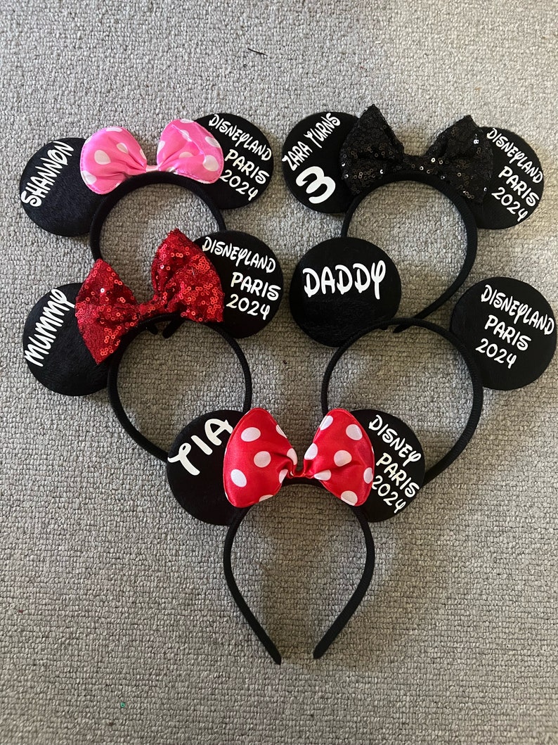 May include: Set of six black mouse ears headbands with white text and colorful bows. The text on the headbands reads "Mummy", "Shannon", "Daddy", "Tara Turns 3", "Yia", and "Disneyland Paris 2024".