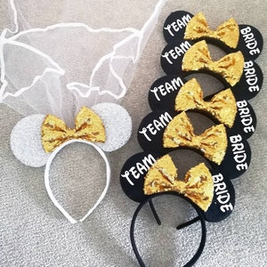 Op de afbeelding: Set van vijf zwarte hoofdbanden met muisoren, gouden glitter strikken en witte tekst. De tekst luidt "Team Bride" op vier van de hoofdbanden en "Bride" op &eacute;&eacute;n hoofdband. Een witte hoofdband met muisoren, een zilveren glitter oor en een gouden glitter strik is ook inbegrepen.