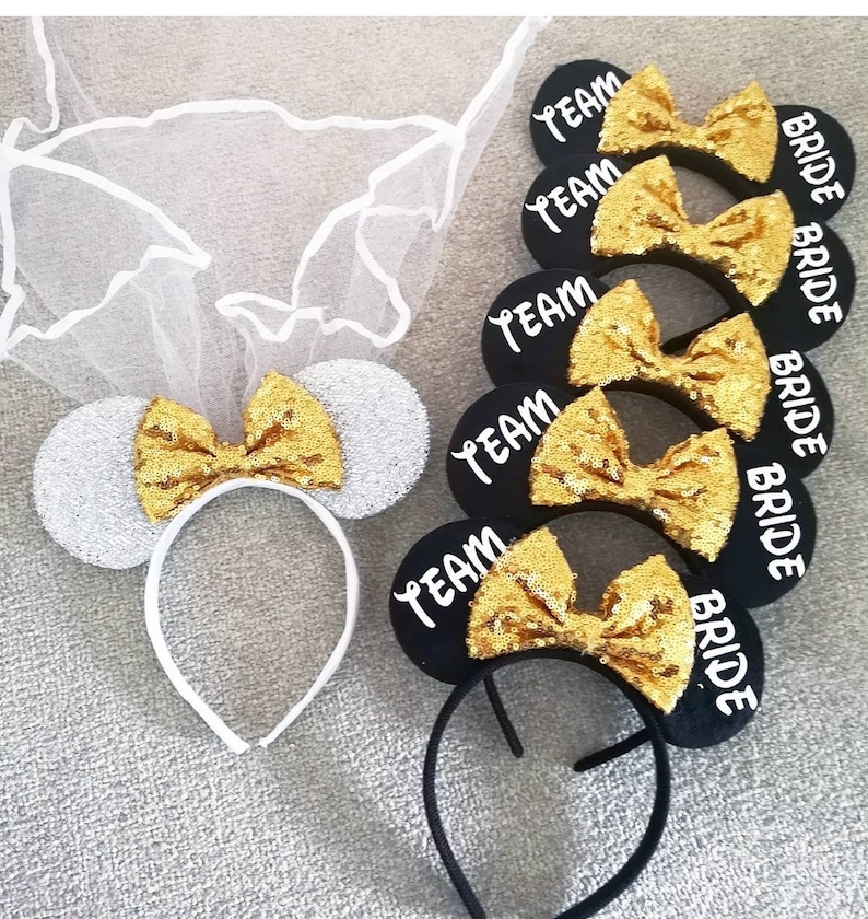 Op de afbeelding: Set van vijf zwarte hoofdbanden met muisoren, gouden glitter strikken en witte tekst. De tekst luidt "Team Bride" op vier van de hoofdbanden en "Bride" op &eacute;&eacute;n hoofdband. Een witte hoofdband met muisoren, een zilveren glitter oor en een gouden glitter strik is ook inbegrepen.