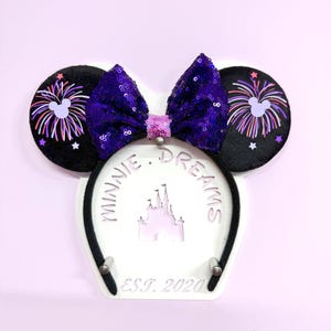 May include: Black Minnie Mouse ears with a purple and pink bow and a firework design on the ears. The ears are on a white stand with the text "Minnie Dreams Est. 2020" and a silhouette of Cinderella's castle.